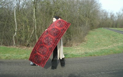 Emine Cerkinaj staat in een straatje, achter haar zien we een veld en bomen. Ze houdt een tapijt voor haar vast. 